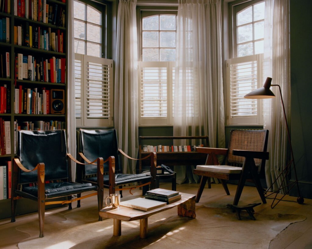 A cozy reading room with tall windows, bookshelves filled with books, wooden chairs, an amber pan on a low table beside books and mugs, and a floor lamp. Sunlight streams softly through sheer curtains.
