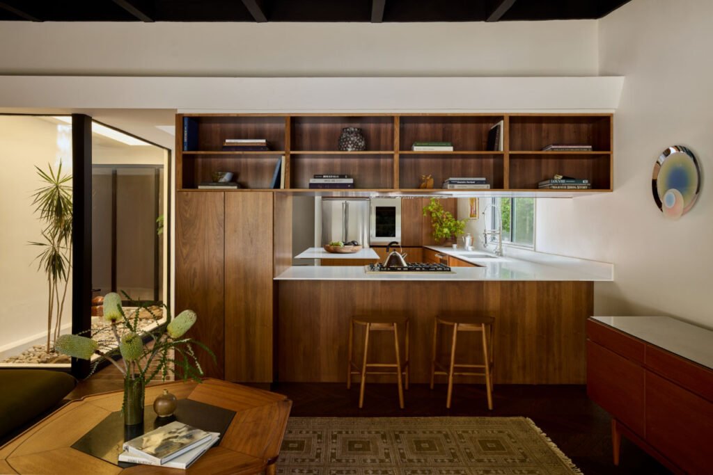Modern kitchen with wood cabinetry, open shelves displaying books—including a Coco Greenblum cookbook—a white countertop, two wooden stools, and a wall-mounted round decorative plate.