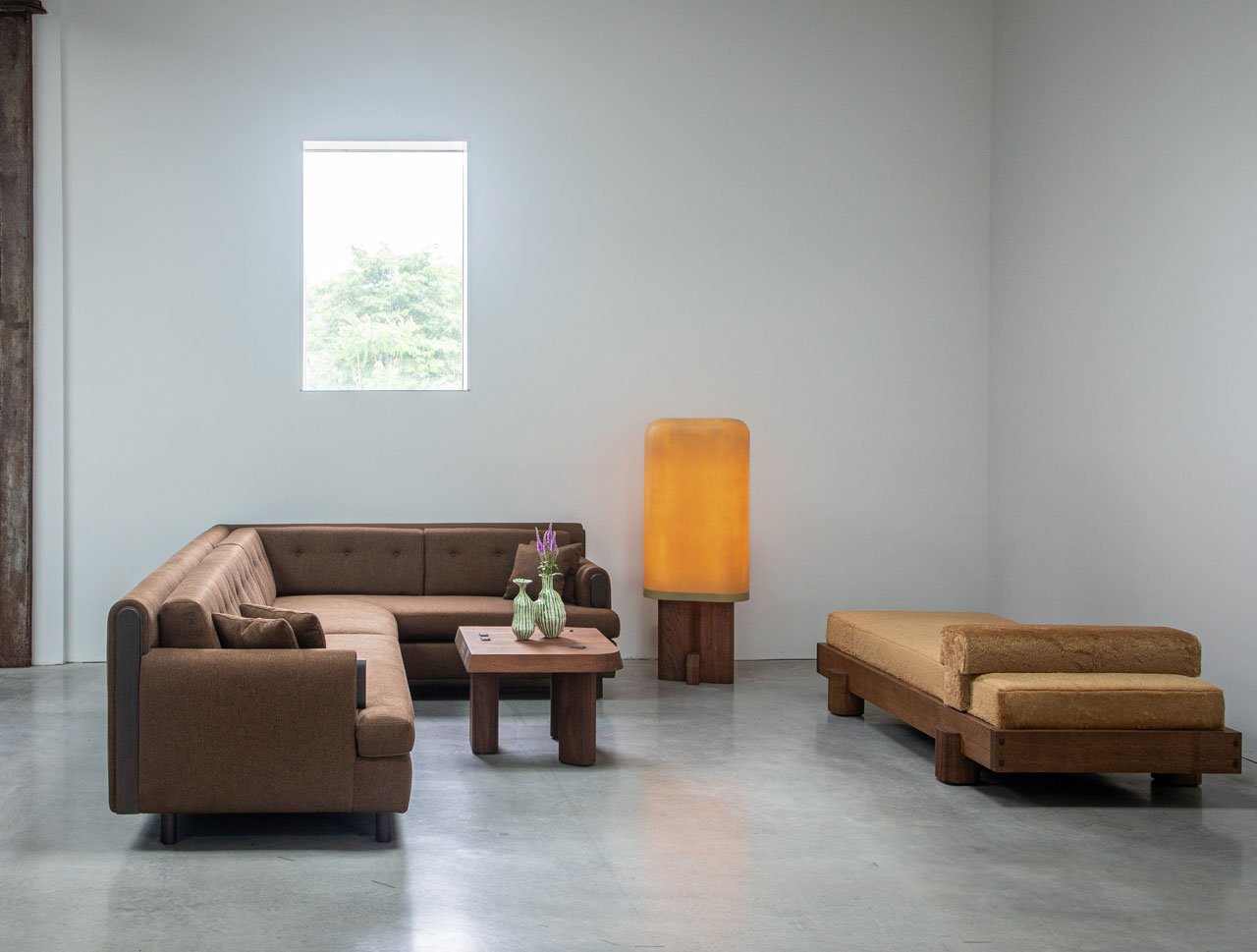 Minimalist living room with a brown sectional sofa, wooden coffee table, large glowing floor lamp, daybed, and a small window on a plain white wall—perfect inspiration for your next Friday Five.