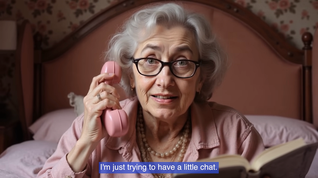 A still from O2’s campaign video showing Daisy, an AI-generated elderly woman with grey hair and glasses, holding a pink telephone handset. A subtitle at the bottom reads “I’m just trying to have a little chat.” She appears warm, slightly confused, and entirely convincing.