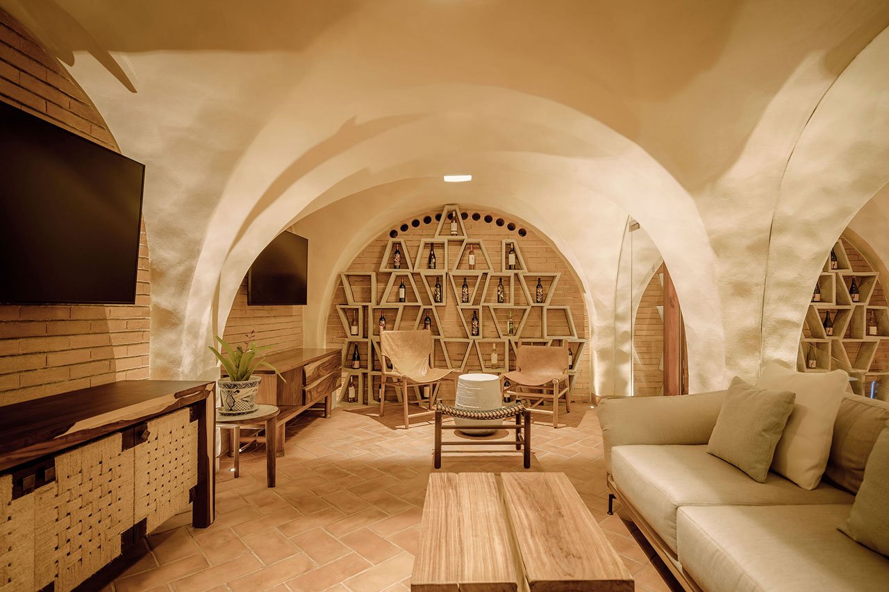 A cozy room with arched ceilings, triangular wooden bookshelves, two chairs, a modern sofa, two flat-screen TVs, and wooden tables on a brick floor.