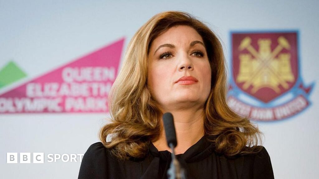 Karren Brady, with long brown hair down to her shoulders and wearing a black dress, standing in front of a microphone