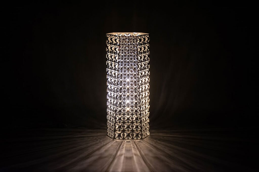 A cylindrical lamp with an intricate, patterned metal shade emits light in a dark room, casting decorative shadows on the surface around it.