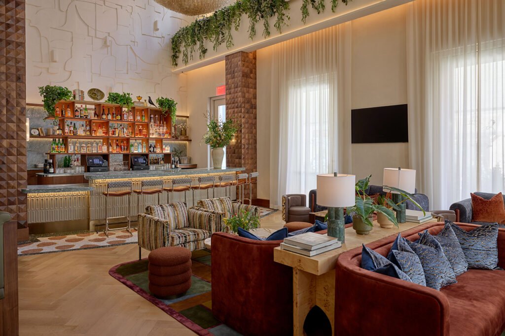 Modern hotel lounge designed by LEGEARD STUDIO, featuring a stylish bar, comfy sofas and armchairs, large windows with sheer curtains, and lush greenery decor on walls and shelves.
