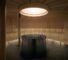 A modern circular sauna with wooden benches lining the walls, a central metal stove filled with rocks, and a gong hanging on the right. Natural light enters from a round ceiling window.
