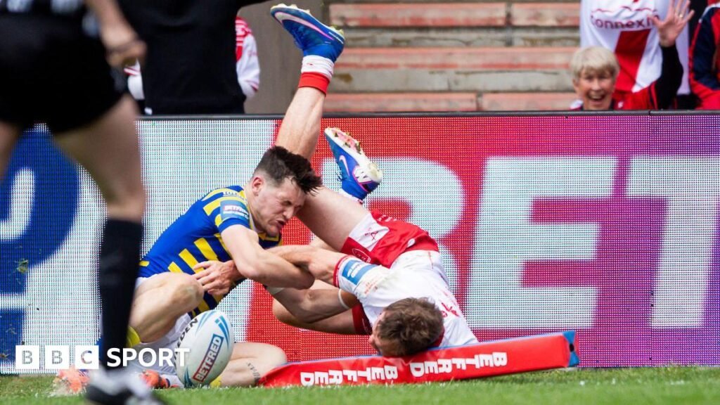 Hull KR player Joe Burgess, in white shirt and red shorts, flattens the corner post as he dives, legs in the air, after scoring a try as Warrington's Josh Thewlis, in blue and yellow hoops, tries to grab him.