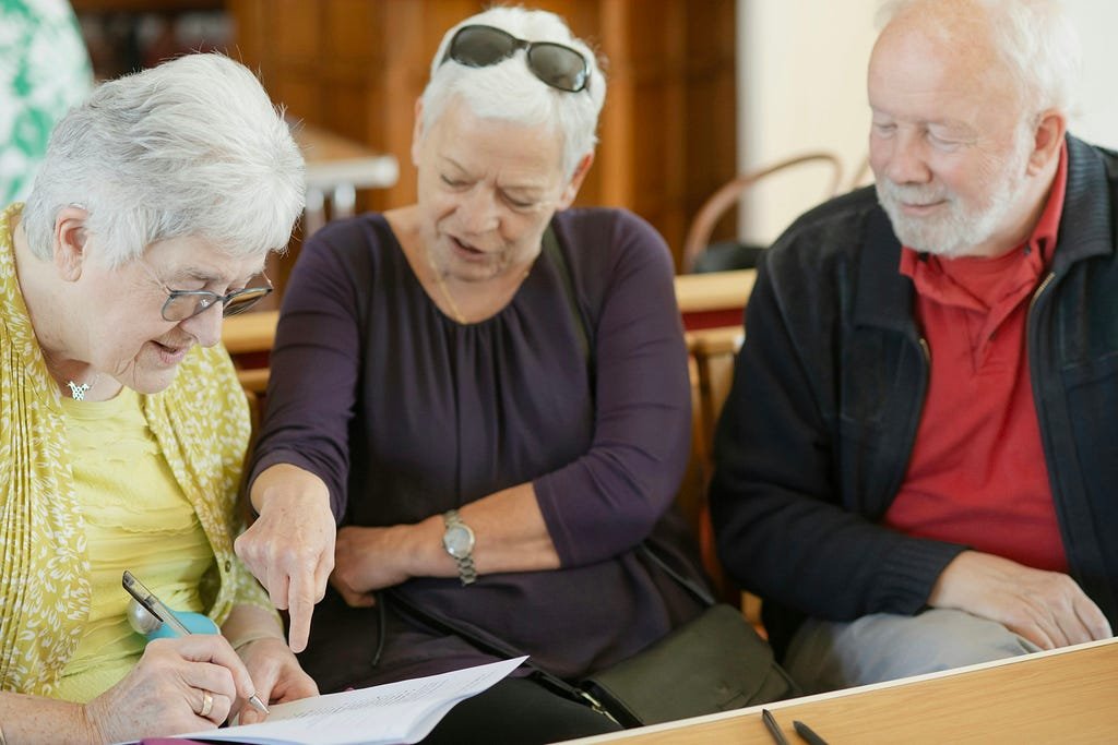 Three older adults sit together at a table reviewing paperwork. One woman points to a document while another writes on it, as a man watches and smiles nearby.