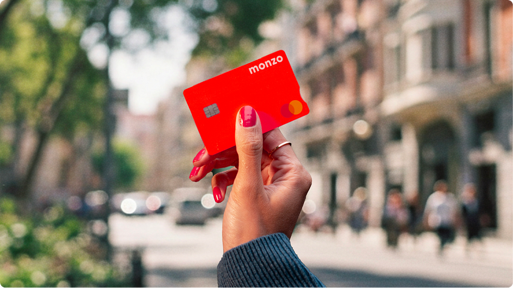 A hand holding up a bright coral Monzo debit card on a sunlit street, with shop fronts and pedestrians blurred in the background.