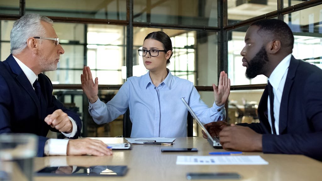 A conflict mediator tries to settle a dispute between two people sitting at a table across from eachother.