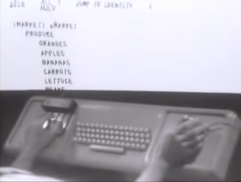 Frame from the 9 December 1968 NLS demonstration. Upper half shows a CRT display with the header “JUMP TO IDENTITY” above an indented hierarchical list reading MARKET, PRODUCE, ORANGES, APPLES, BANANAS, CARROTS, LETTUCE, BEANS. Lower half shows the Engelbart workstation: left hand on the five-key chord keyset, right hand on the three-button mouse, standard keyboard between them