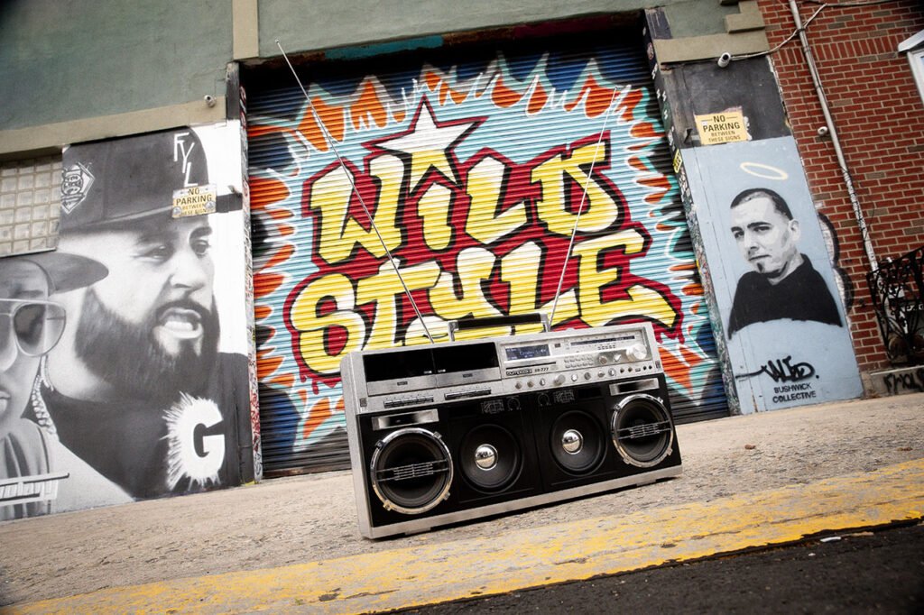 A large boombox sits on the ground in front of a mural reading "WILD STYLE," surrounded by black-and-white portraits and graffiti art on a city wall.