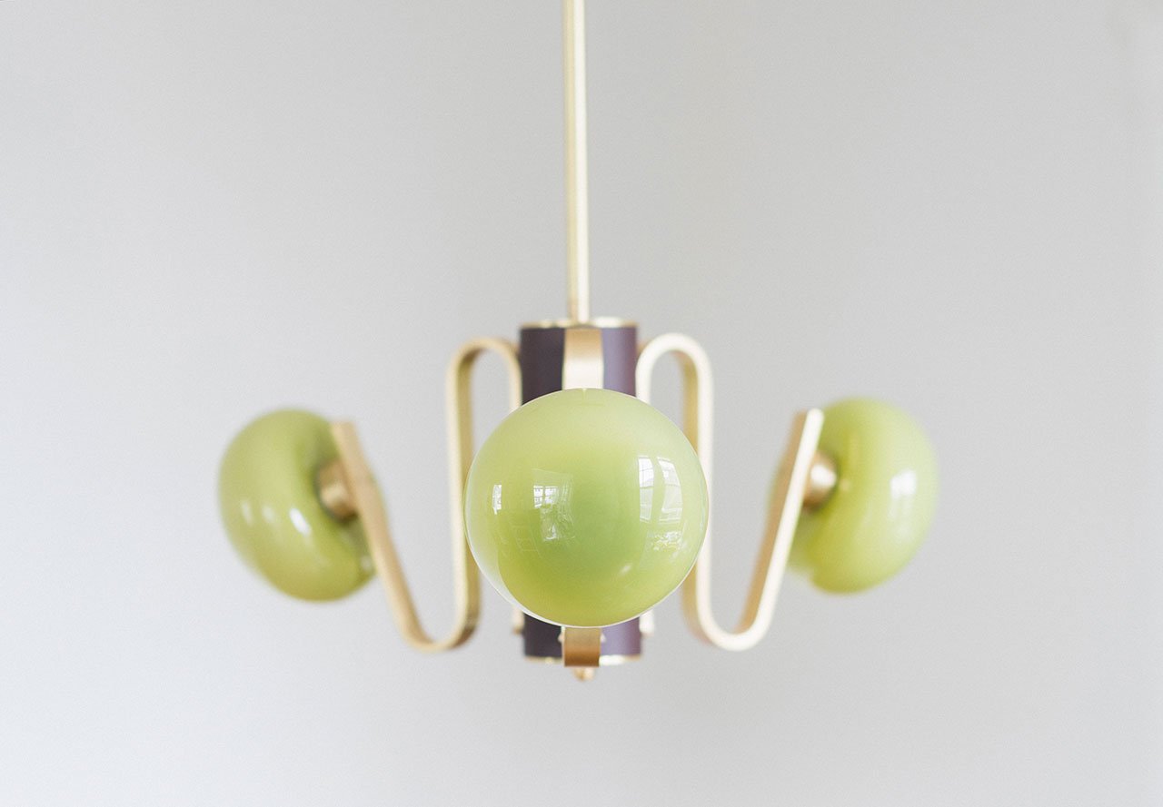 A modern chandelier by Sara Schoenberger, featuring three spherical green glass shades and gold metal arms, suspended against a plain light gray background.