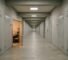 A long institutional corridor with identical numbered doors, one standing open to reveal a warmly lit room with a desk and bookshelf. All other doors are closed.