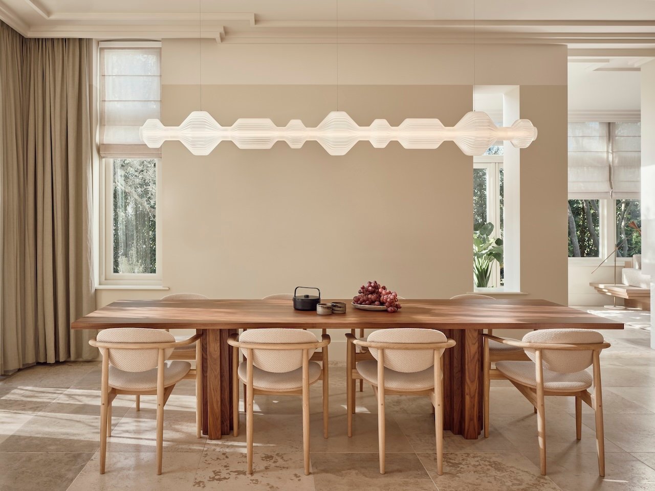 A modern dining room with a long wooden table, six upholstered chairs, a sculptural pendant light, and minimal table decor. Large windows let in natural light.