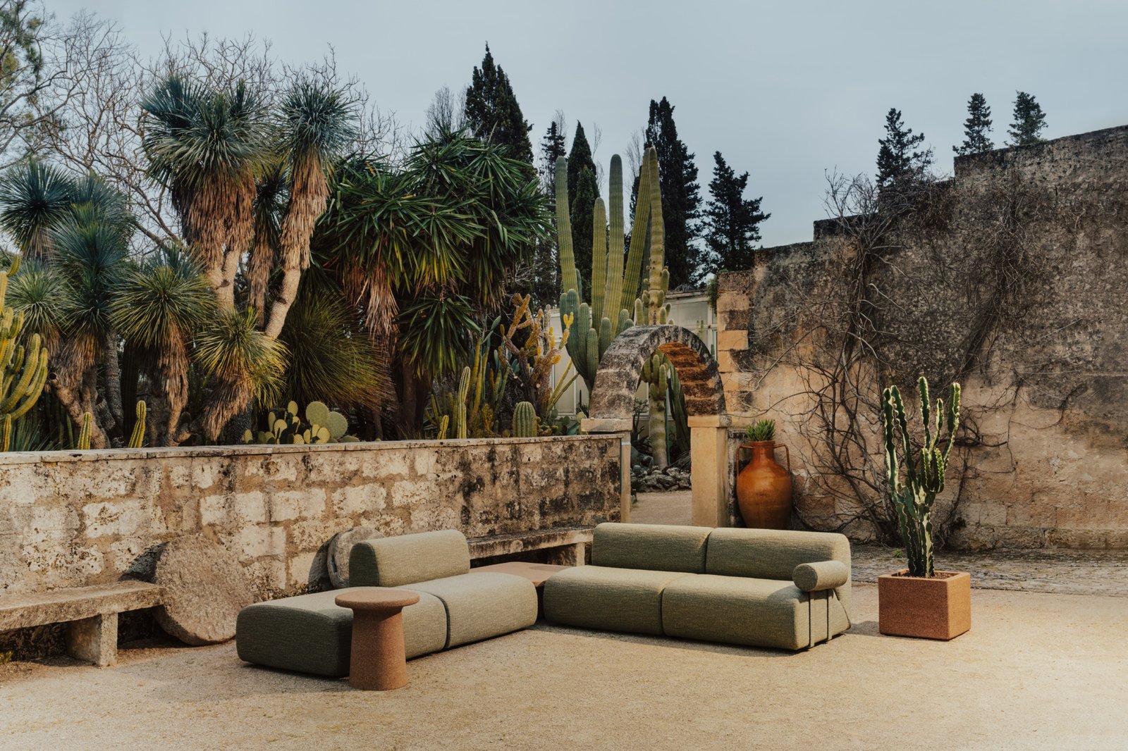 Outdoor seating area with modular sofas, a side table, and potted cacti, set against a stone wall and surrounded by various desert plants and trees.
