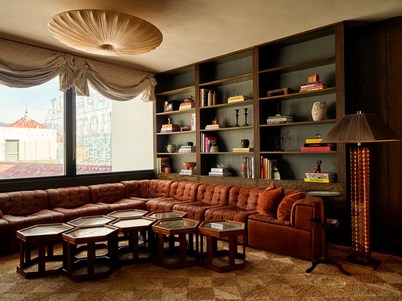 A cozy living room with a brown leather sectional, hexagonal coffee tables, built-in shelves with books and decor, and a large window with draped curtains.