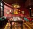 A restaurant dining area designed by Nicole Alexander of Siren Betty features pink chairs, set tables, teal barstools, a decorative chandelier, and art on red paneled walls. Large windows let in natural light.