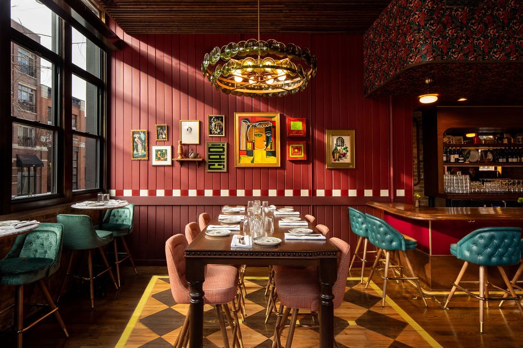 A restaurant dining area designed by Nicole Alexander of Siren Betty features pink chairs, set tables, teal barstools, a decorative chandelier, and art on red paneled walls. Large windows let in natural light.
