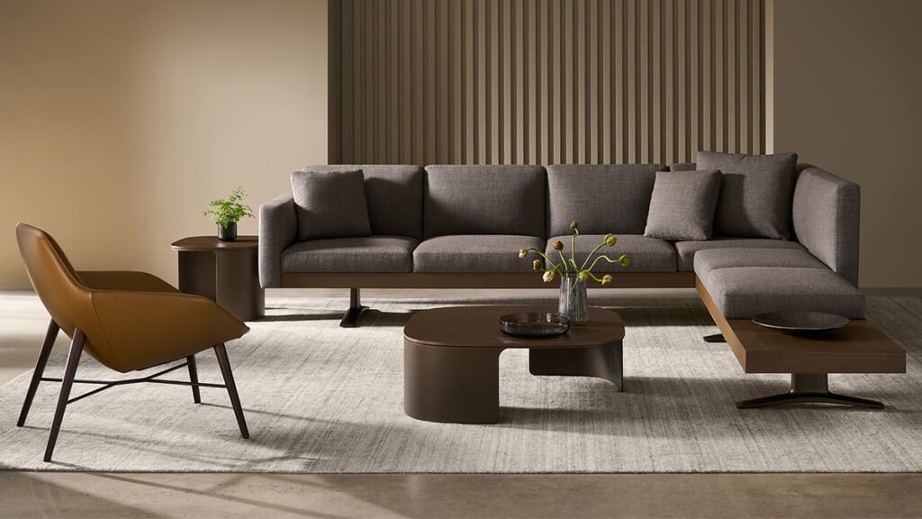 Modern living room with a gray sectional sofa, brown accent chair, wooden coffee tables, and neutral walls. Plants and decorative objects sit on tables atop a light rug.