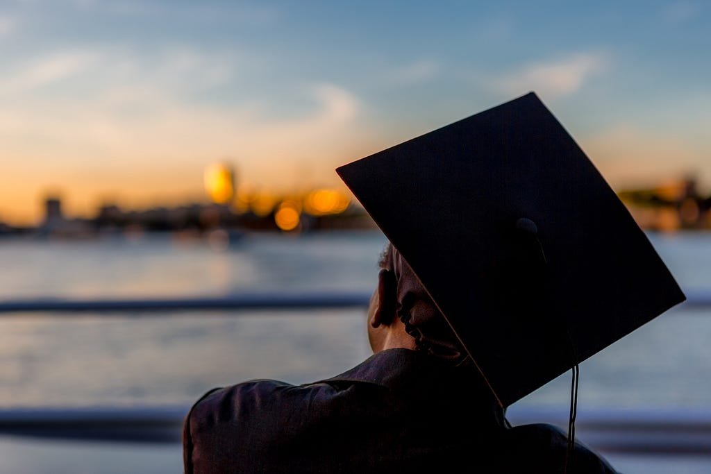 A graduate hat rests on the ground near a serene body of water, symbolizing achievement and reflection.