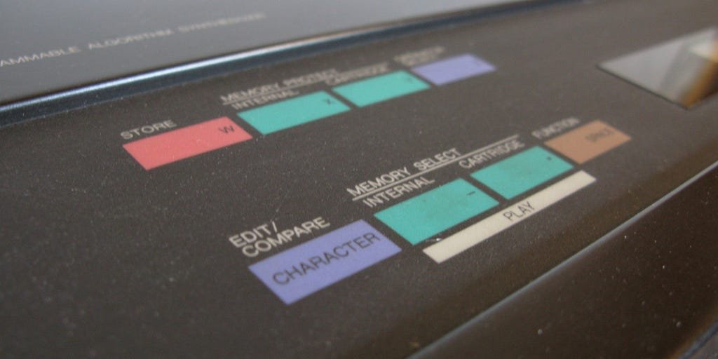 Close-up of a Yamaha DX7 synthesizer panel: pastel teal, pink and lavender buttons labelled Store, Memory select, Internal, Cartridge and Play, on a black case.